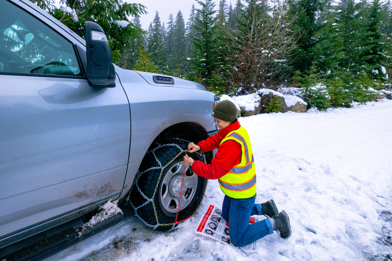 A lady installing a tire chain on a pickup truck. She is kneeling in the snow and is fastening the chain to the Les Schwab Open Range A/T Plus tire.